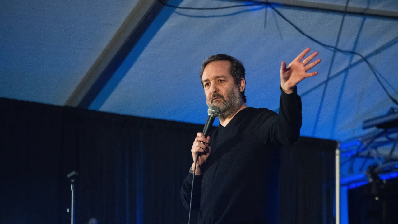 Comedian with a hand raised mid-story under blue stage lights